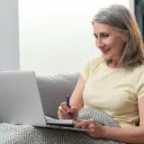 Mujer mayor sonríe mientras toma notas en un cuaderno, quizás repasando las fases de la memoria en su ordenador portátil.