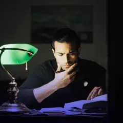 Hombre concentrado bajo una lámpara verde, buscando cómo memorizar un tema en un libro.