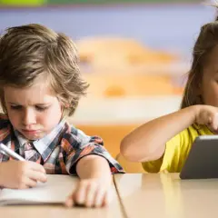 Niño con expresión de frustración escribiendo en un cuaderno, mientras una niña ríe usando una tablet. Contraste entre la educación tradicional y la moderna.