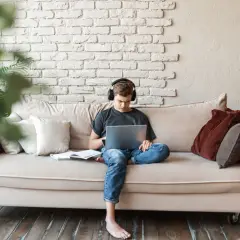 Joven con auriculares, concentrado en su portátil, sentado en un sofá con libros. Un espacio cómodo para estudiar en la biblioteca de casa.