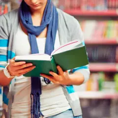 Chica con bufanda azul lee un libro verde en una biblioteca, decidida a estudiar más.