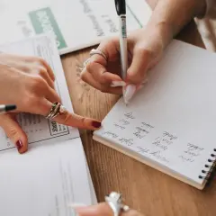 Manos escribiendo en un cuaderno y señalando un examen, una forma de estudiar para un examen de desarrollo.