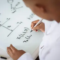 Un niño con bata blanca escribe fórmulas en una pizarra, aprendiendo trucos para resolver problemas de primaria.
