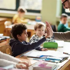 Un maestro da un "choca esos cinco" a un niño en clase, celebrando el aprendizaje y educando emociones. Otros niños observan sonrientes.