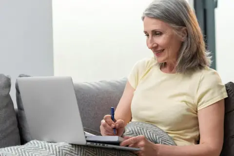 Mujer mayor sonríe mientras toma notas en un cuaderno, quizás repasando las fases de la memoria en su ordenador portátil.
