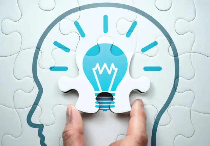 Manos ensamblan un rompecabezas que forma una cabeza con una bombilla de idea, simbolizando el aprendizaje cognitivo y la chispa de la inspiración.