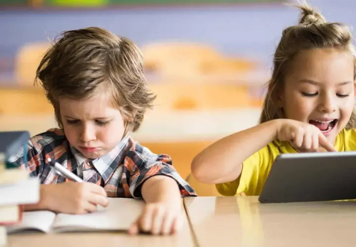 Niño con expresión de frustración escribiendo en un cuaderno, mientras una niña ríe usando una tablet. Contraste entre la educación tradicional y la moderna.
