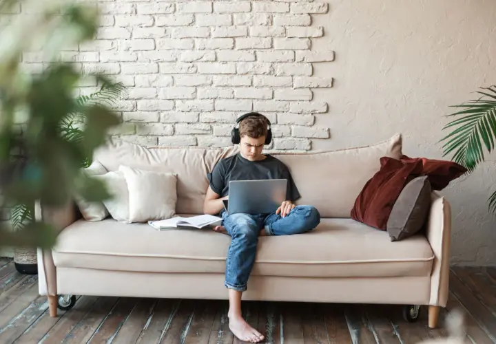 Joven con auriculares, concentrado en su portátil, sentado en un sofá con libros. Un espacio cómodo para estudiar en la biblioteca de casa.