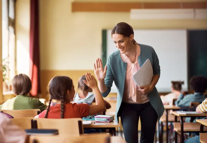 Maestra sonríe y da un cinco a una alumna, fomentando la educación personalizada en un aula con otros niños.
