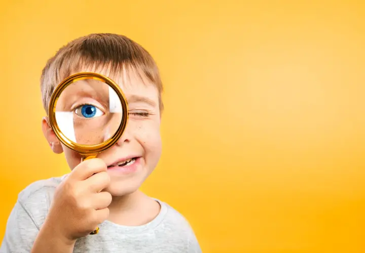 Niño con pecas mira a través de lupa dorada, ojo azul brillante. Un ejemplo de pedagogía activa, explorando el mundo con curiosidad.