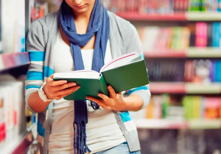 Chica con bufanda azul lee un libro verde en una biblioteca, decidida a estudiar más.