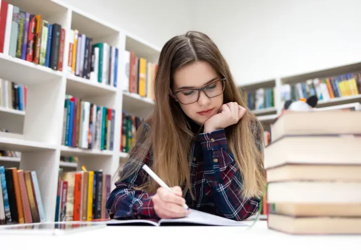 Joven concentrada en su estudio biblioteca en casa, rodeada de estanterías llenas de libros.