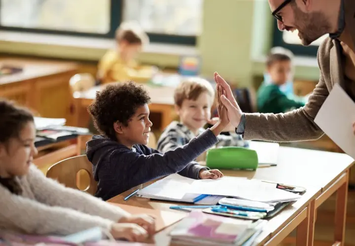 Un maestro da un "choca esos cinco" a un niño en clase, celebrando el aprendizaje y educando emociones. Otros niños observan sonrientes.