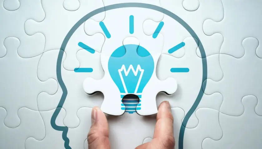 Manos ensamblan un rompecabezas que forma una cabeza con una bombilla de idea, simbolizando el aprendizaje cognitivo y la chispa de la inspiración.