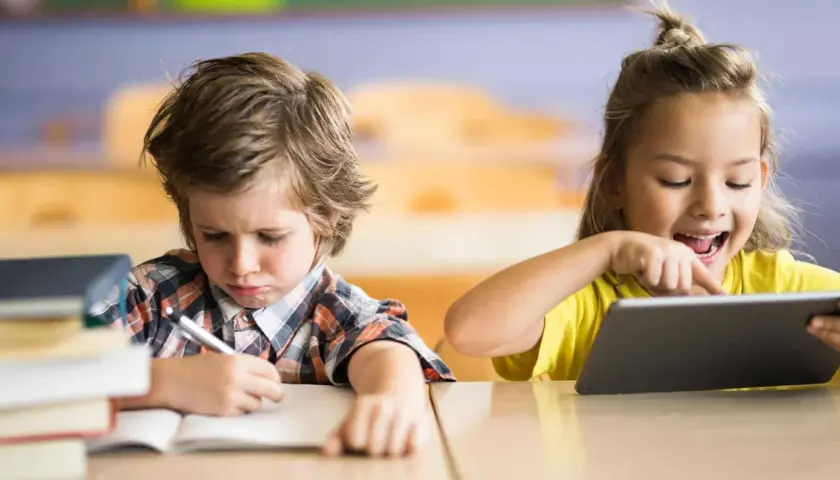 Niño con expresión de frustración escribiendo en un cuaderno, mientras una niña ríe usando una tablet. Contraste entre la educación tradicional y la moderna.