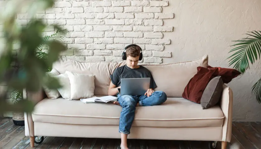 Joven con auriculares, concentrado en su portátil, sentado en un sofá con libros. Un espacio cómodo para estudiar en la biblioteca de casa.