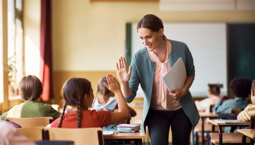 Maestra sonríe y da un cinco a una alumna, fomentando la educación personalizada en un aula con otros niños.