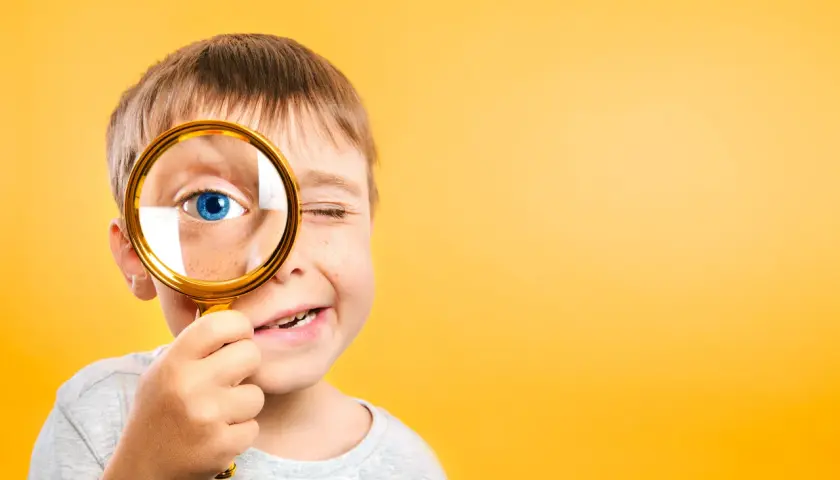 Niño con pecas mira a través de lupa dorada, ojo azul brillante. Un ejemplo de pedagogía activa, explorando el mundo con curiosidad.