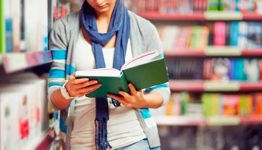 Chica con bufanda azul lee un libro verde en una biblioteca, decidida a estudiar más.