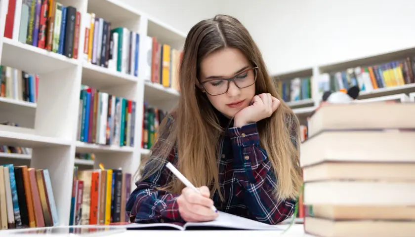 Joven concentrada en su estudio biblioteca en casa, rodeada de estanterías llenas de libros.
