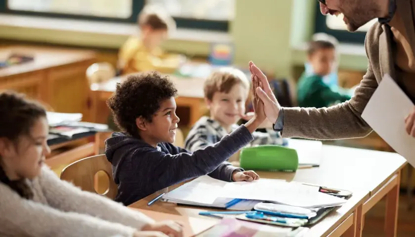 Un maestro da un "choca esos cinco" a un niño en clase, celebrando el aprendizaje y educando emociones. Otros niños observan sonrientes.