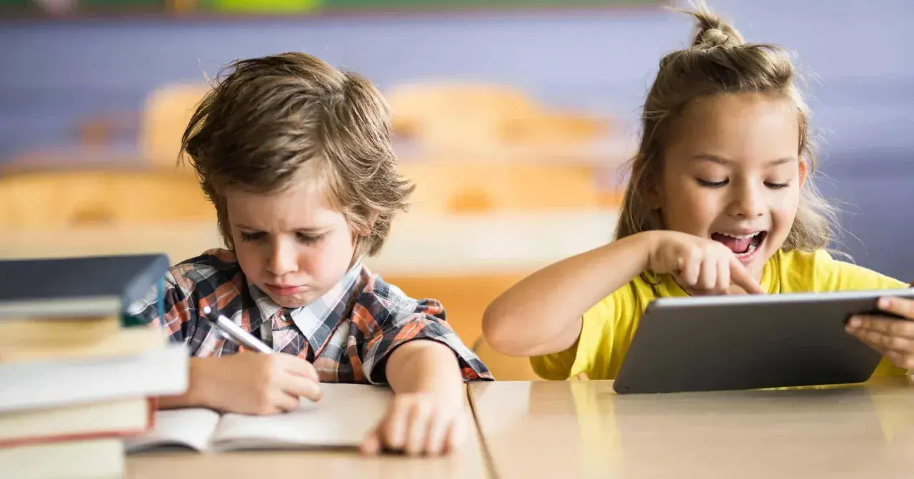 Niño con expresión de frustración escribiendo en un cuaderno, mientras una niña ríe usando una tablet. Contraste entre la educación tradicional y la moderna.