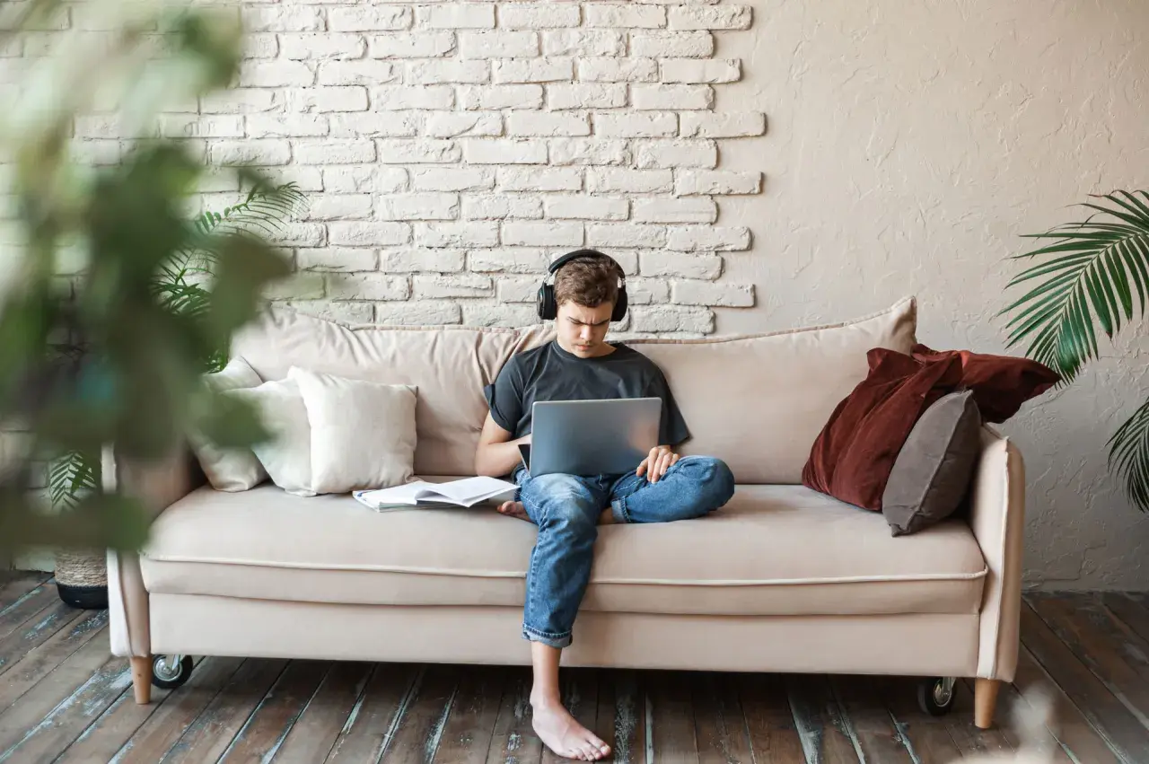 Joven con auriculares, concentrado en su portátil, sentado en un sofá con libros. Un espacio cómodo para estudiar en la biblioteca de casa.