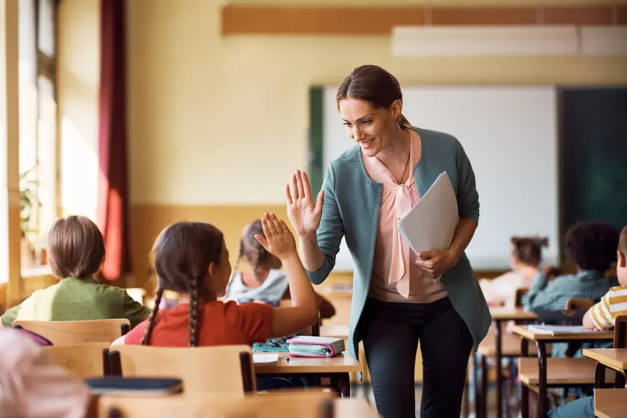 Maestra sonríe y da un cinco a una alumna, fomentando la educación personalizada en un aula con otros niños.