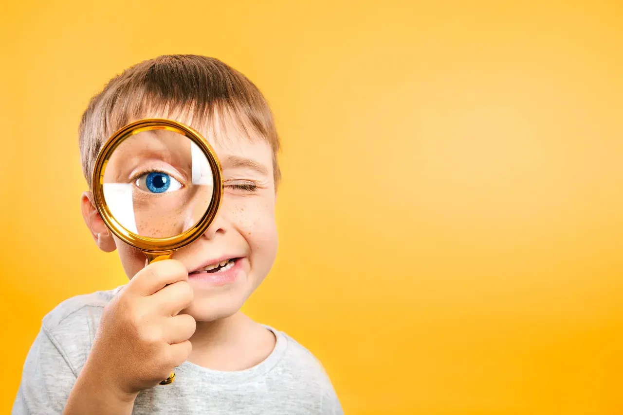 Niño con pecas mira a través de lupa dorada, ojo azul brillante. Un ejemplo de pedagogía activa, explorando el mundo con curiosidad.