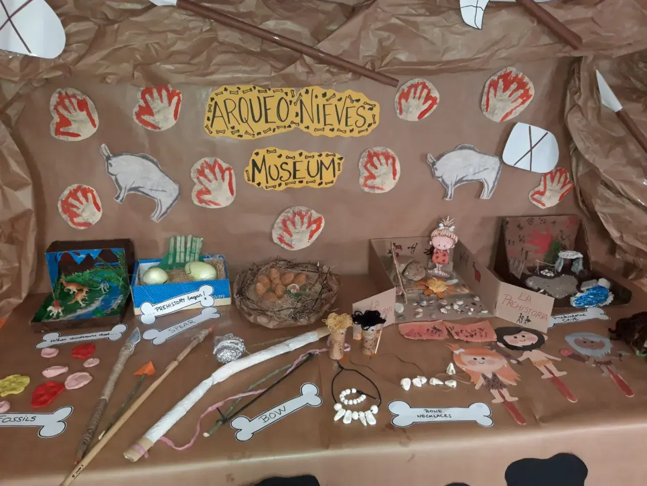 Exhibición de trabajo por proyectos infantil sobre la prehistoria: fósiles, lanzas, arcos, collares de huesos y figuras de hombres de las cavernas.