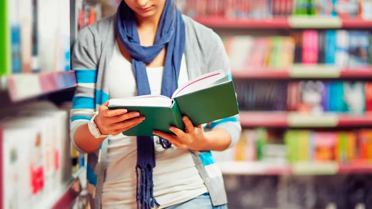 Chica con bufanda azul lee un libro verde en una biblioteca, decidida a estudiar más.