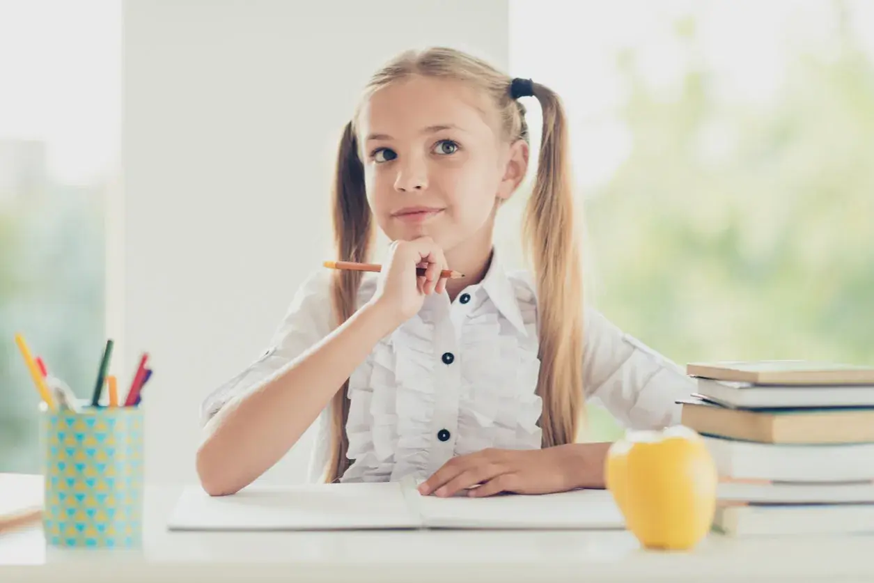 Niña pensativa con lápiz, rodeada de útiles escolares y libros. Ideas para **técnicas de estudio para niños**.