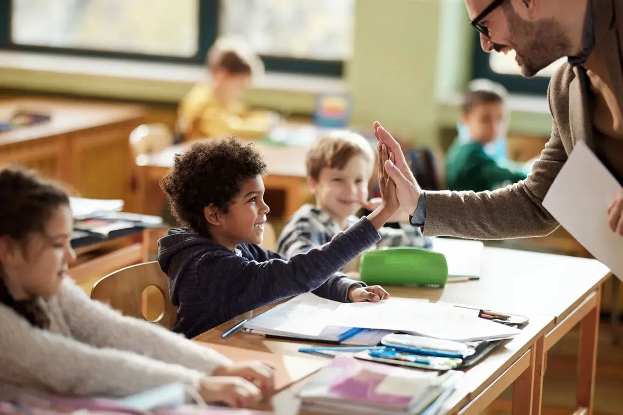 Un maestro da un "choca esos cinco" a un niño en clase, celebrando el aprendizaje y educando emociones. Otros niños observan sonrientes.