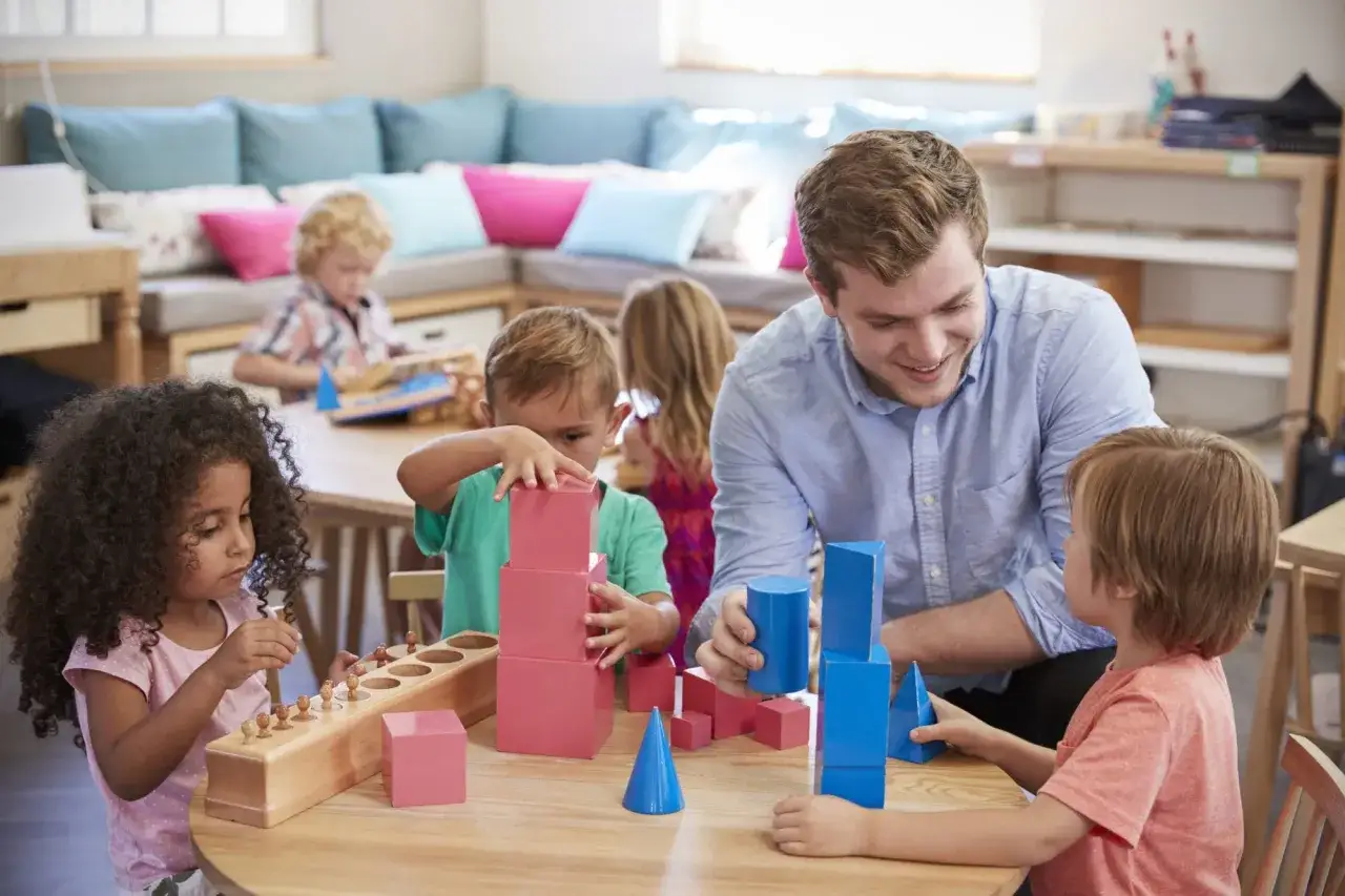Un hombre sonriente ayuda a ni&ntilde;os a construir con bloques de colores, fomentando el desarrollo cognitivo en un aula.