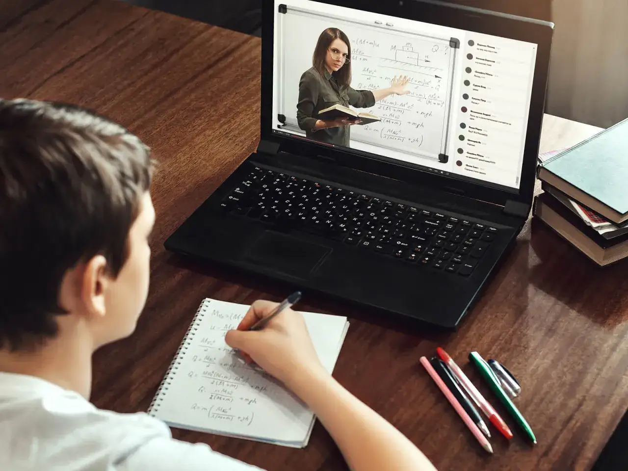 Un niño estudia en línea, tomando notas mientras una profesora explica física en la pantalla.