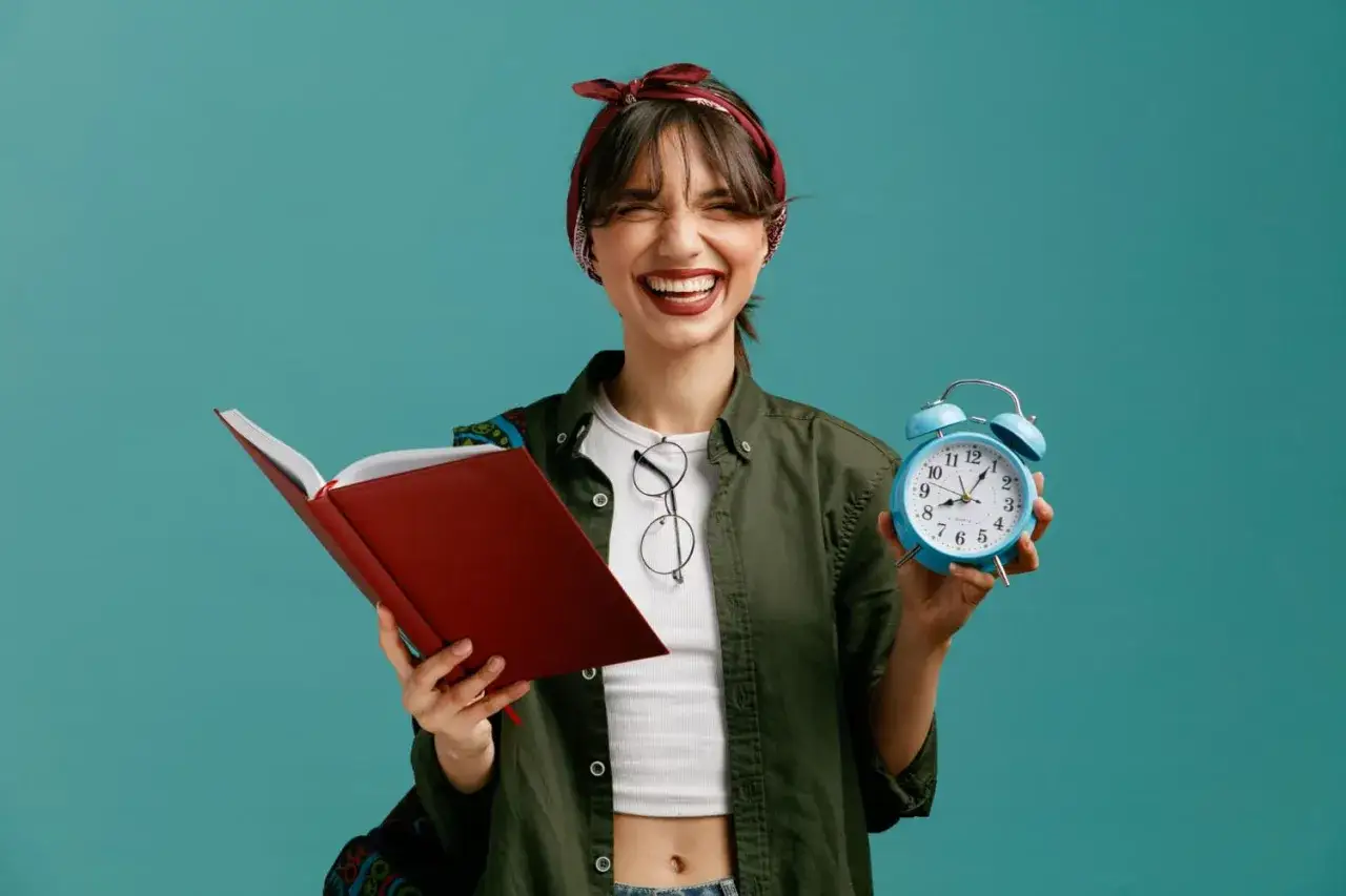 Mujer sonriente con libro rojo y despertador azul, lista para aplicar el metodo flowtime y optimizar su tiempo de estudio.