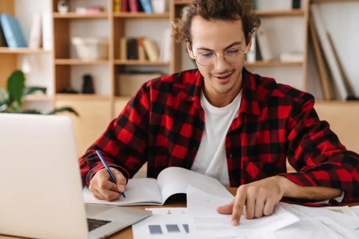 Joven con camisa a cuadros rojos y negros, frente a una laptop, escribe en un cuaderno. A pesar de que me cuesta retener información, se esfuerza por aprender.