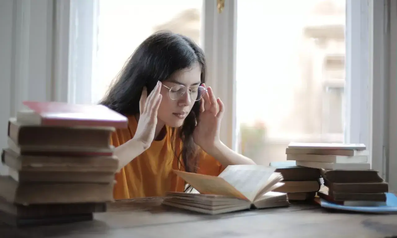 Joven con gafas, rodeada de libros, se toca las sienes. Reflexiona sobre qué hacer antes de un examen, buscando concentración.