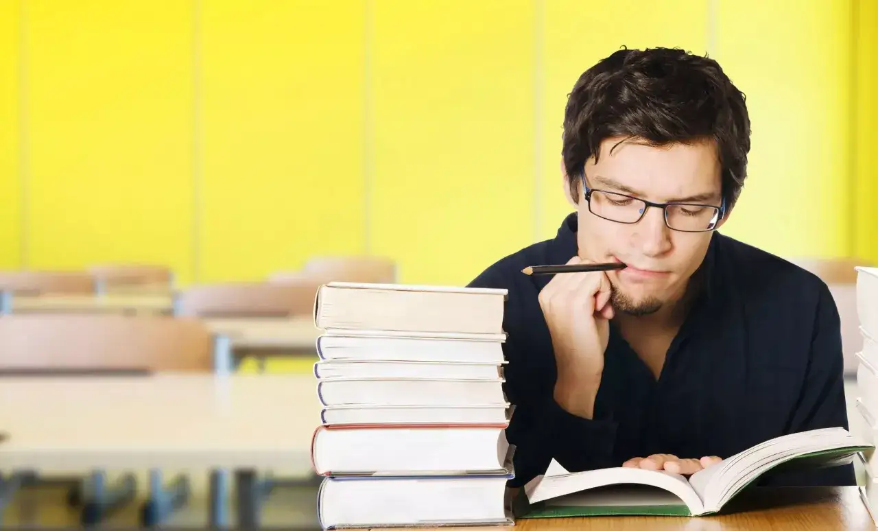 Estudiante con gafas y lápiz en la boca, rodeado de libros, pensando cómo memorizar para un examen.
